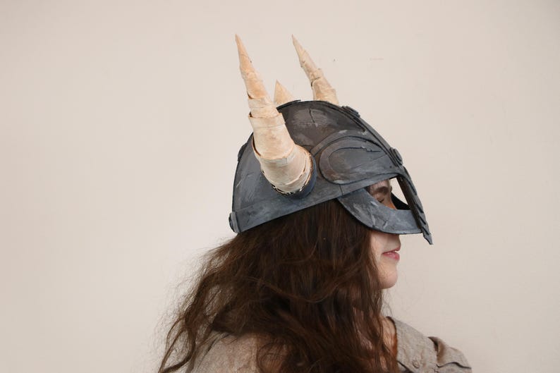 May include: A gray Viking helmet with two large, cream-colored horns. The helmet has a dark gray finish and is worn by a person with long, brown hair. The helmet is a costume piece.