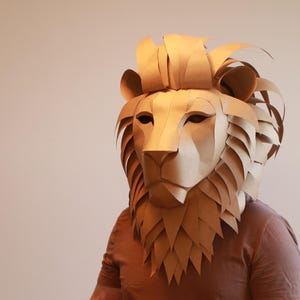 Op de afbeelding: Een bruin kartonnen leeuwenmasker met een gedetailleerde manen en gezicht. Het masker wordt gedragen door een persoon, maar het gezicht van de persoon is niet zichtbaar.