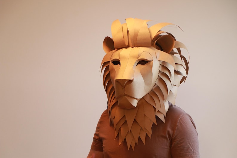 Op de afbeelding: Een bruin kartonnen leeuwenmasker met een gedetailleerde manen en gezicht. Het masker wordt gedragen door een persoon, maar het gezicht van de persoon is niet zichtbaar.