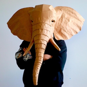 Puede incluir: Una gran máscara de elefante hecha a mano con papel marrón. La máscara presenta pliegues y arrugas detalladas, con orejas grandes y una larga trompa. La persona que lleva la máscara viste una chaqueta negra.