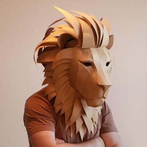 Op de afbeelding: Een persoon draagt ​​een bruin papier-mach&eacute; leeuwenmasker. Het masker is gedetailleerd met ingewikkelde vouwen en kreuken, waardoor het een realistisch uiterlijk krijgt.