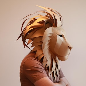 Op de afbeelding: Een persoon draagt ​​een bruin papier-mach&eacute; leeuwenmasker. Het masker is gedetailleerd met een manen van papieren stroken en een realistisch leeuwen gezicht.
