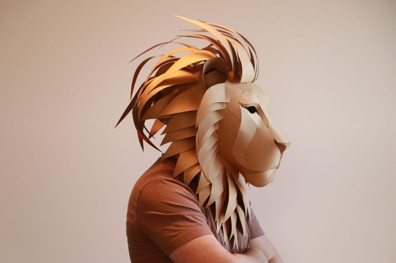 Op de afbeelding: Een persoon draagt ​​een bruin papier-mach&eacute; leeuwenmasker. Het masker is gedetailleerd met een manen van papieren stroken en een realistisch leeuwen gezicht.