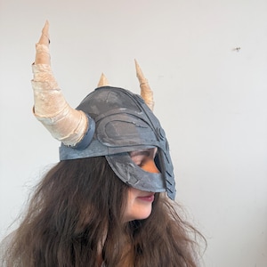 May include: A gray helmet with a face guard and four tan horns. The helmet is worn by a person with long brown hair. The horns are made of a rolled material. The helmet is a costume piece.