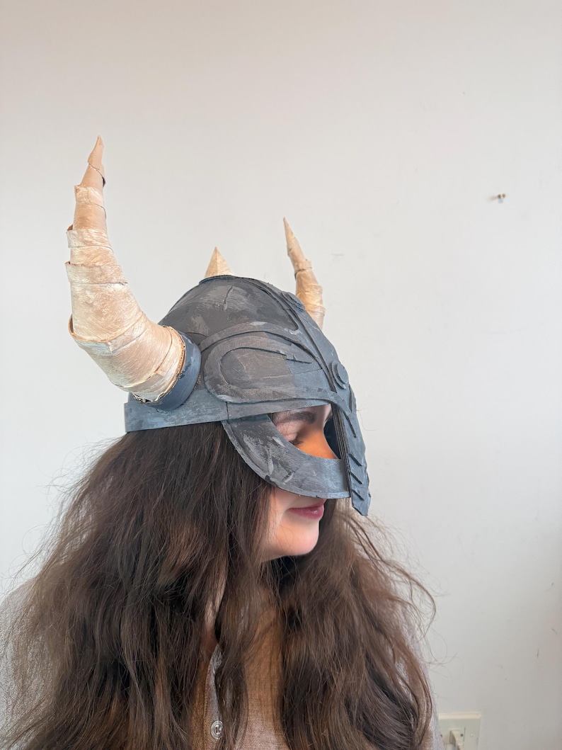 May include: A gray helmet with a face guard and four tan horns. The helmet is worn by a person with long brown hair. The horns are made of a rolled material. The helmet is a costume piece.