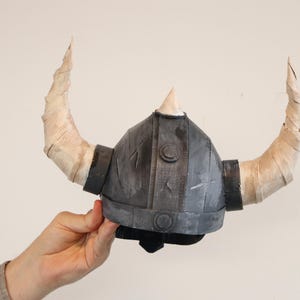 May include: A gray and black Viking helmet with two large, curved, tan horns. The helmet has a pointed top and is held by a person's hand. The horns are wrapped in a light-colored material.
