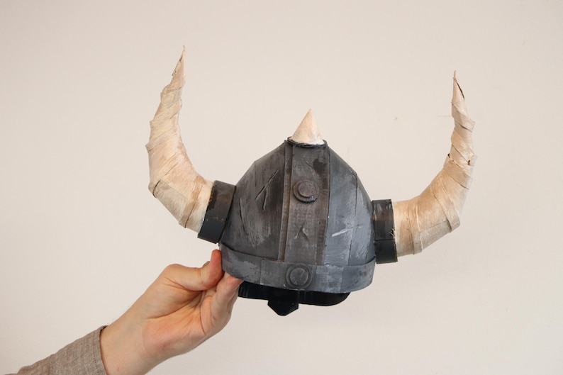 May include: A gray and black Viking helmet with two large, curved, tan horns. The helmet has a pointed top and is held by a person's hand. The horns are wrapped in a light-colored material.