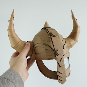 May include: A handmade cardboard Viking helmet with large, curved horns. The helmet is constructed from layered cardboard pieces, featuring decorative circular accents and a pointed top. The horns are made from rolled cardboard.