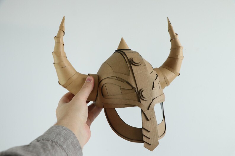 May include: A handmade cardboard Viking helmet with large, curved horns. The helmet is constructed from layered cardboard pieces, featuring decorative circular accents and a pointed top. The horns are made from rolled cardboard.