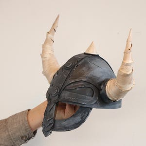 May include: A gray Viking helmet with two large, curved horns and a smaller central horn. The helmet is held up by a person's arm. The horns are wrapped in a light-colored material.