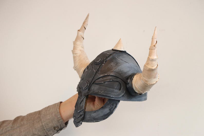 May include: A gray Viking helmet with two large, curved horns and a smaller central horn. The helmet is held up by a person's arm. The horns are wrapped in a light-colored material.