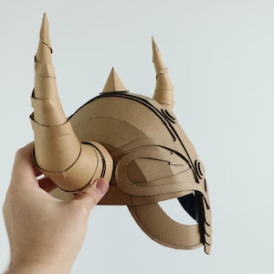 May include: A brown cardboard helmet with two large, curved horns and a pointed crest. The helmet has intricate cut-out details and is held against a white background.