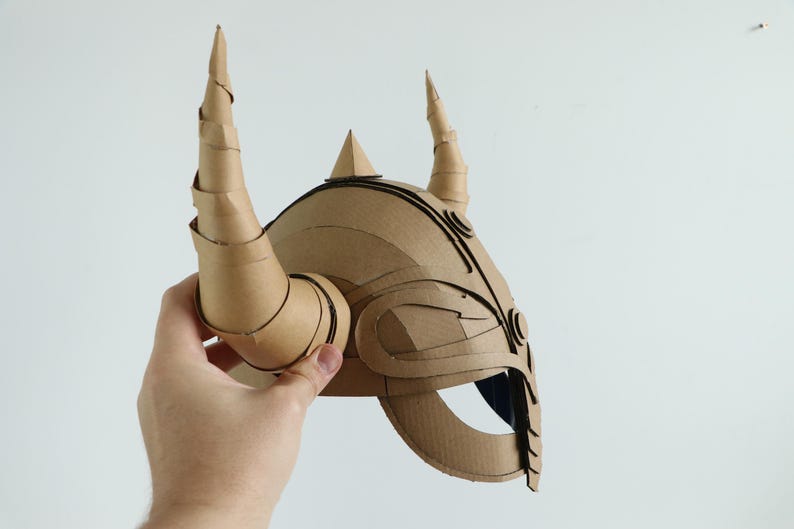 May include: A brown cardboard helmet with two large, curved horns and a pointed crest. The helmet has intricate cut-out details and is held against a white background.