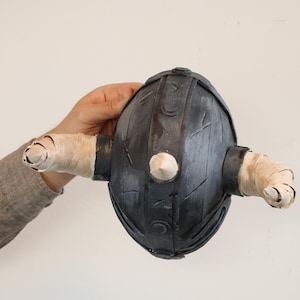 May include: A dark gray, egg-shaped object with two cylindrical protrusions wrapped in white tape. A white, conical shape is centered on the front. The object is held against a plain white background.