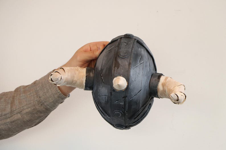 May include: A dark gray, egg-shaped object with two cylindrical protrusions wrapped in white tape. A white, conical shape is centered on the front. The object is held against a plain white background.