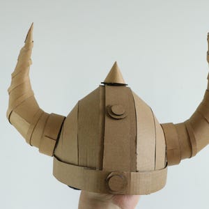 May include: A handmade Viking helmet crafted from corrugated cardboard. The helmet features a rounded crown, a central conical spike, and two large, curved horns. The cardboard is a natural brown color, with visible seams and construction details.