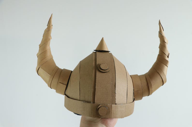 May include: A handmade Viking helmet crafted from corrugated cardboard. The helmet features a rounded crown, a central conical spike, and two large, curved horns. The cardboard is a natural brown color, with visible seams and construction details.