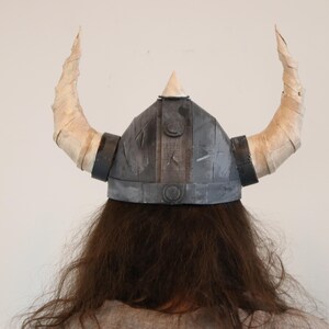 May include: A gray Viking helmet with two large, curved, cream-colored horns. The helmet has a conical top and is worn over long, brown hair. The horns are wrapped in a textured material, and the helmet has black bands.