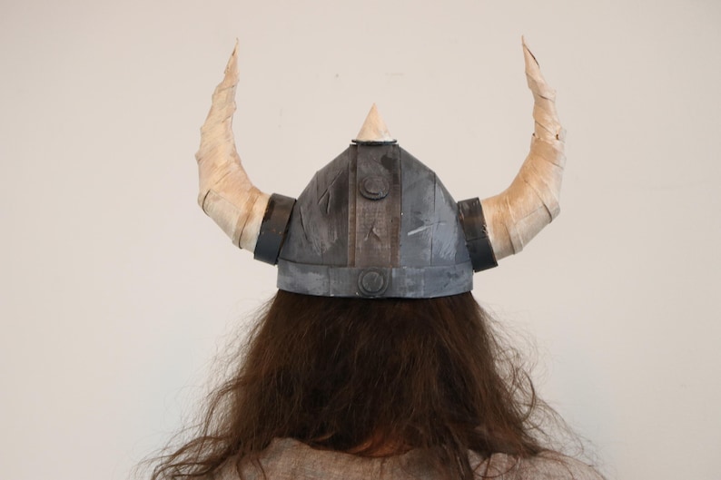 May include: A gray Viking helmet with two large, curved, cream-colored horns. The helmet has a conical top and is worn over long, brown hair. The horns are wrapped in a textured material, and the helmet has black bands.