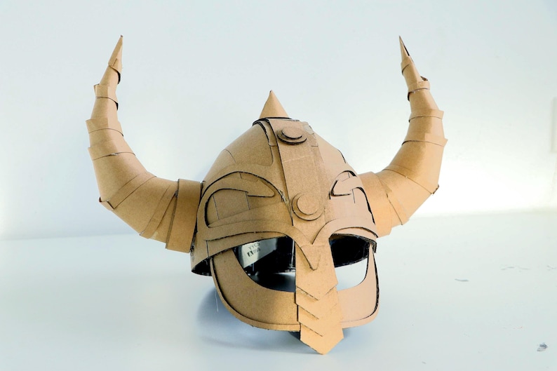 May include: A handmade cardboard Viking helmet with large, curved horns. The helmet is constructed from corrugated cardboard, with visible seams and edges. The design includes a pointed top and a face guard, showcasing a DIY craft project.
