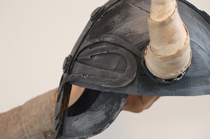 May include: A gray Viking helmet with a curved design and a single horn. The helmet has a distressed, textured appearance with visible brushstrokes. The horn is a light beige color.