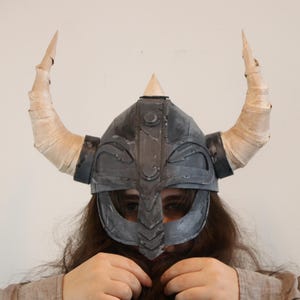 May include: A dark gray Viking helmet with two large, curved, cream-colored horns. The helmet has a triangular peak and a face guard. The horns are wrapped in a light-colored material. The helmet is a costume piece.