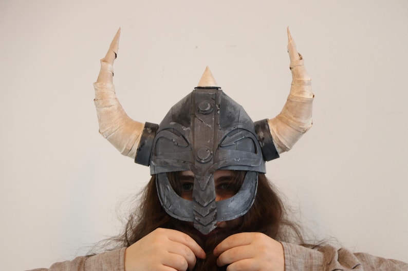 May include: A dark gray Viking helmet with two large, curved, cream-colored horns. The helmet has a triangular peak and a face guard. The horns are wrapped in a light-colored material. The helmet is a costume piece.
