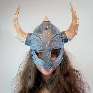 May include: A gray and black Viking helmet with two large, curved, tan horns. The helmet has a detailed design with rivets and a pointed top. The horns are wrapped in a textured material, creating a rustic appearance.