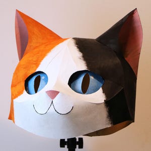 May include: A colorful cat mask with orange, black, and white fur. The cat has large blue eyes, a pink nose, and a smiling mouth. The mask is likely made of paper or cardboard and is designed to be worn.