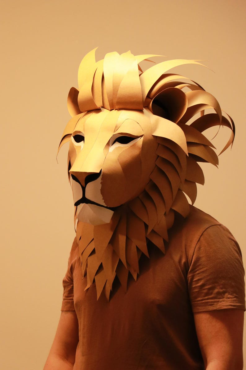 Op de afbeelding: Een persoon draagt ​​een bruin papieren mach&eacute;-leeuwenmasker. Het masker heeft een gedetailleerde manen en realistische gezichtskenmerken.