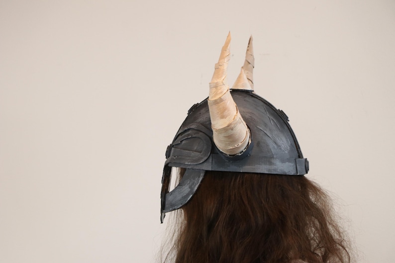 May include: A gray Viking helmet with two large, curved, cream-colored horns. The helmet has a dark gray face guard and is worn over long, brown hair. The helmet is a costume piece.