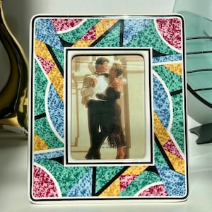 May include: A square ceramic picture frame with a colorful geometric pattern in teal, yellow, pink, and blue. The frame holds a photograph of a couple embracing. The frame has a black border and a white background.