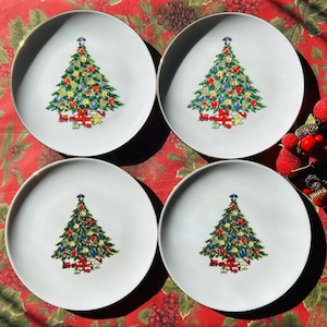 May include: Four white ceramic plates with a gold rim, each featuring a colorful Christmas tree design. The trees are decorated with ornaments and sit above wrapped gifts. The plates are arranged on a red floral tablecloth.