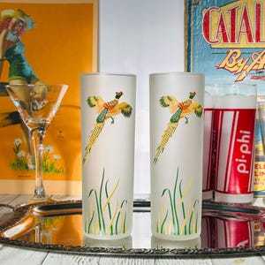 May include: Two frosted highball glasses with a pheasant design. The glasses have a white background with a colorful pheasant and green and yellow grass. The glasses are on a silver tray.