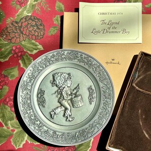 May include: A silver-colored Hallmark plate from 1978, featuring a raised relief of the Little Drummer Boy. The plate has a floral border and is accompanied by a box and a card that reads "Christmas 1978 The Legend of the Little Drummer Boy."