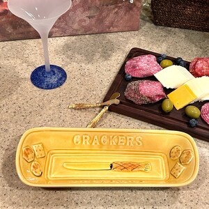 May include: A yellow ceramic cracker dish with the word "CRACKERS" printed on it. The dish has a knife design and a small section for crackers.