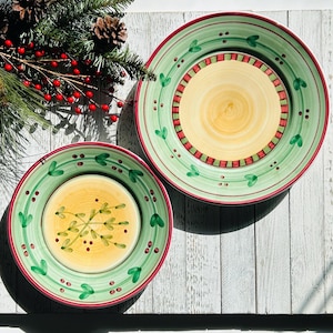 May include: Two decorative ceramic plates with a green border, red trim, and a yellow center. The plates have a festive design with green leaves and red berries. A sprig of pine and red berries is in the background.