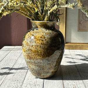 May include: A glazed ceramic vase with a speckled brown and gold finish, holding dried flowers. The vase has a rounded shape and a wide opening. The background includes a framed picture and a decorative metal piece.