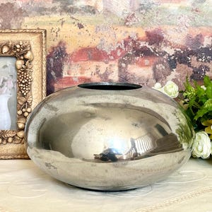 May include: A large, silver-toned, oval-shaped vase with a wide opening. The vase reflects the surrounding environment. A framed picture and white flowers are visible in the background. The vase is a decorative home decor item.