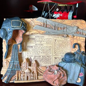 May include: A decorative wall plaque featuring a vintage travel theme. The plaque includes a miniature airplane, a pilot's helmet, a model of the Eiffel Tower, and luggage. The plaque is made of a textured material and has a brown and beige color scheme. The text on the plaque reads "MAHOGANY, 30000 FT. on Mahogany boards, plank, each is perfectly seasoned, and the best part of it is that it is made by WM. FISK, Washington, D.C. at the Military Yard, and of equal quality."