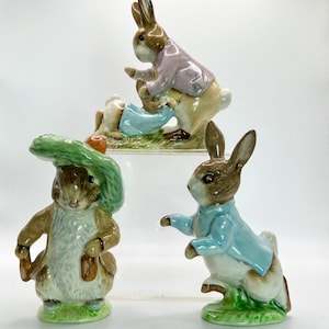 May include: Three ceramic rabbit figurines. One rabbit wears a green hat and holds a bag. Another rabbit wears a blue jacket and points. The third figurine depicts two rabbits interacting, one in a pink jacket.