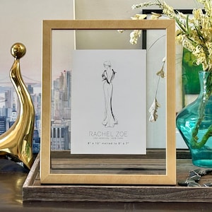 May include: A gold-framed picture with a fashion illustration by Rachel Zoe, matted to 12.7cm x 17.8cm. A gold sculpture and a turquoise vase with flowers are also visible. The scene is set on a wooden tray.