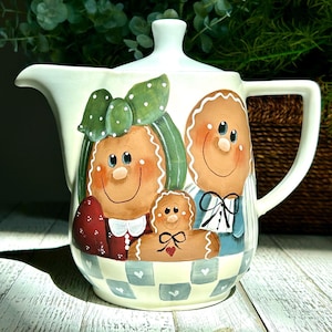 May include: White ceramic jug with a painted design of a gingerbread family. The family consists of a mother, father, and a small child. The mother is wearing a green bow and a red dress. The father is wearing a blue shirt and a black bow tie. The child is wearing a red bow. The family is sitting on a checkered tablecloth.