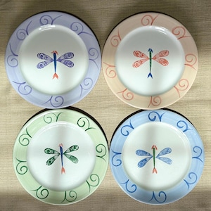 May include: Four ceramic plates featuring a dragonfly design. Each plate has a white centre with a dragonfly and a coloured border with decorative swirls. Colours include lavender, peach, green, and blue.