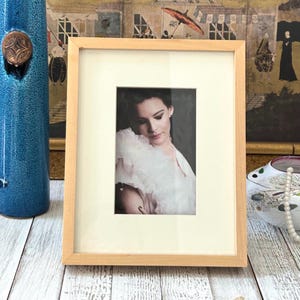 May include: A rectangular, light-colored wooden picture frame with a cream-colored mat. The frame holds a portrait of a woman wearing a white ruffled garment. A blue vase and decorative items are in the background.