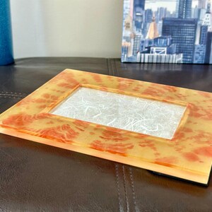 Op de afbeelding: Een rechthoekige fotolijst met een heldere, getextureerde harsafwerking. Het frame heeft een geel en oranje gevlekt patroon. Het midden van het frame heeft een wit, vezelig materiaal. Het frame ligt op een donkerbruin oppervlak.