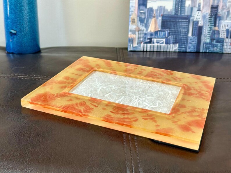 Op de afbeelding: Een rechthoekige fotolijst met een heldere, getextureerde harsafwerking. Het frame heeft een geel en oranje gevlekt patroon. Het midden van het frame heeft een wit, vezelig materiaal. Het frame ligt op een donkerbruin oppervlak.
