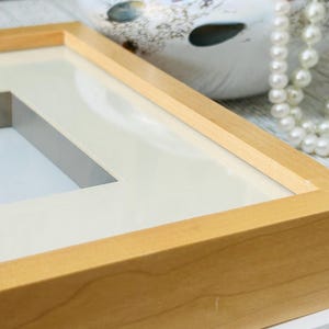 May include: Close-up of a light-colored wooden picture frame with a white mat. The frame has a rectangular opening and is constructed with corner joints. A pearl necklace and a decorative ceramic object are in the background.