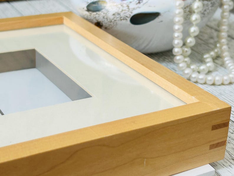 May include: Close-up of a light-colored wooden picture frame with a white mat. The frame has a rectangular opening and is constructed with corner joints. A pearl necklace and a decorative ceramic object are in the background.