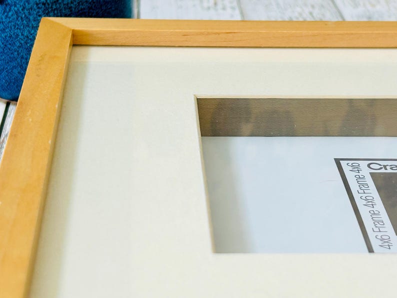 May include: A close-up of a wooden picture frame with a cream-colored mat. The frame is light brown and has a rectangular opening for a 4x6 inch photo. The frame is on a white surface.